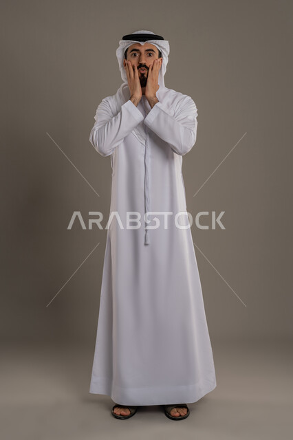 النظر إلى الكاميرا بإيماءات الذهول ,الانزعاج عند سماع خبر سيء، التفاجئ من شيء ما، الشعور بالصدمة والاستغراب , بورتريه لرجل عربي خليجي اماراتي يرتدي الزي التقليدي والغترة و يرفع يديه على وجهه بتعابير الدهشة, صورة الجسم بالكامل، خلفية رمادية