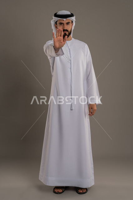 طلب التوقف عن فعل شيء ما، الأمر بالوقوف وعدم التمادي، الرفض وعدم الخضوع، النهي عن التقدم وأهمية ترك مسافة أمان، بورتريه لرجل عربي خليجي اماراتي يرتدي الكندورة والغترة يرفع كفه للأعلى، الجسم بالطول الكامل، خلفية رمادية