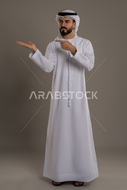 Containment and pointing with index finger to a hologram element, portrait of an Arab Emirati Gulf man wearing a kandura and ghutra raising his hand to display a hologram element, gestures indicating concentration, full body, gray background