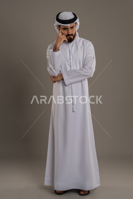 Gestures of contemplation, reflection and concentration, portrait of an Arab Gulf Emirati man wearing a kandura and ghutra, standing and putting his hand under his chin, looking somewhere with an expression of thought, planning to solve a problem, feeling confused and uncertain, full body, gray background