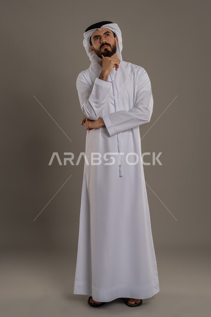 Gestures of contemplation, reflection and concentration, portrait of an Arab Gulf Emirati man wearing a kandura and ghutra, standing and putting his hand under his chin, looking somewhere with an expression of thought, planning to solve a problem, feeling confused and uncertain, full body, gray background