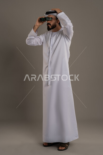 Planning and setting future goals, using binoculars to see distant objects, clarifying and zooming in, portrait of an Arab Gulf Emirati man wearing a kandura and ghutra standing and putting a telescope with telescope lenses on his eyes and looking somewhere, full body length, gray background