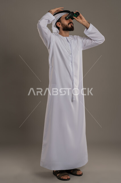 استخدام المنظار ثنائي العينين لمشاهدة الأشياء البعيدة، توضيح الرؤية وتقريبها، التخطيط ووضع الأهداف المستقبلية بورتريه لرجل عربي خليجي اماراتي يرتدي الكندورة والغترة يقف ويضع تلسكوب العدسات المقربة على عينيه وينظر الى الاعلى، الجسم بالطول الكامل، خلفية رمادي