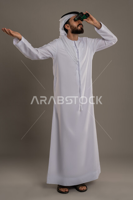 استخدام المنظار ثنائي العينين لمشاهدة الأشياء البعيدة، التخطيط ووضع الأهداف المستقبلية، توضيح الرؤية وتقريبها، بورتريه لرجل عربي خليجي اماراتي يرتدي الكندورة والغترة يقف ويضع تلسكوب العدسات المقربة على عينيه وينظر الى الاعلى، الجسم بالطول الكامل، خلفية رمادي