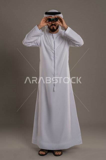 التخطيط ووضع الأهداف المستقبلية، استخدام المنظار ثنائي العينين لمشاهدة الأشياء البعيدة، توضيح الرؤية وتقريبها، بورتريه لرجل عربي خليجي اماراتي يرتدي الكندورة والغترة يقف ويضع تلسكوب العدسات المقربة على عينيه وينظر الى الكاميرا، الجسم بالطول الكامل، خلفية رمادي