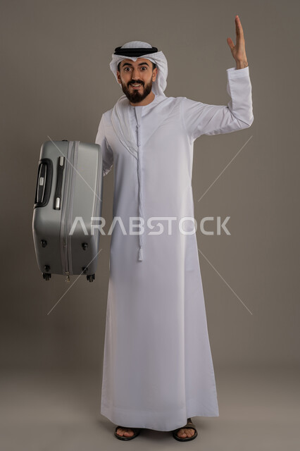 مفهوم السياحة والسفر في إجازة الصيف، الفرح والاستعداد لقضاء رحلة صيفية ممتعة، القيام بجولة ترفيهية، بورتريه لرجل عربي خليجي اماراتي يرتدي الكندورة والغترة يحمل بيده حقيبة الأمتعة ينظر إلى الكاميرا بإيماءات السرور، الجسم بالطول الكامل، خلفية رمادية