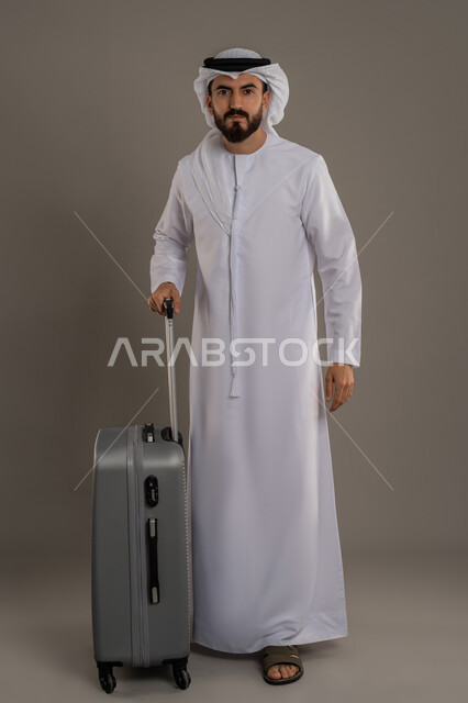 الفرح والاستعداد لقضاء رحلة صيفية ممتعة، القيام بجولة ترفيهية، بورتريه لرجل عربي خليجي اماراتي يرتدي الكندورة والغترة يقف بجانب حقيبة الأمتعة ينظر إلى الكاميرا بإيماءات السرور، مفهوم السياحة والسفر في إجازة الصيف، الجسم بالطول الكامل، خلفية رمادية