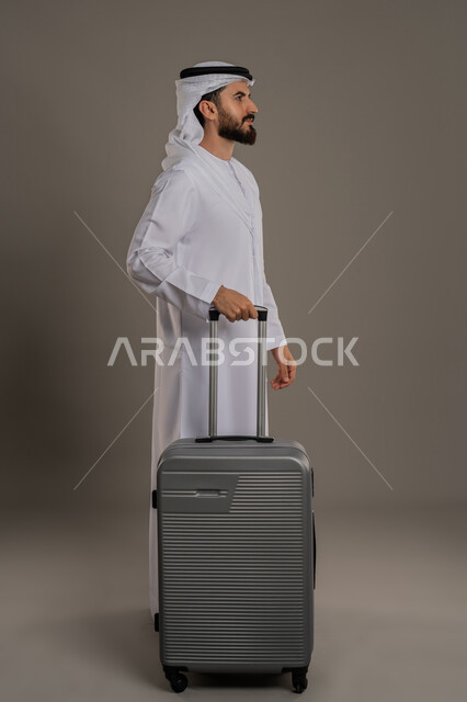 القيام بجولة ترفيهية، بورتريه لرجل عربي خليجي اماراتي يرتدي الكندورة والغترة يقف بجانب حقيبة الأمتعة ينظر إلى مكان ما بإيماءات السرور، الفرح والاستعداد لقضاء رحلة صيفية ممتعة، مفهوم السياحة والسفر في إجازة الصيف، الجسم بالطول الكامل، خلفية رمادية