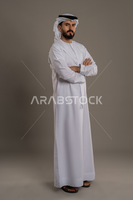 الاهتمام بالأناقة والمظهر الخارجي، بورتريه لرجل عربي خليجي اماراتي يرتدي الكندورة والغترة يقف باستقامة وبايدي متقاطعة، النظر الى الكاميرا بإيماءات الجدية والثقة في النفس، مفهوم الوسامة والرجولة والقوة، الجسم الكامل بالطول، خلفية رمادية