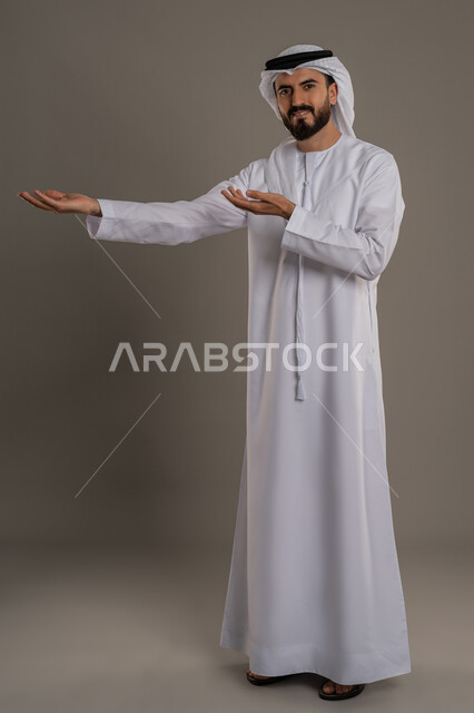 حركات وايماءات تدل على هذا الموقع, الاشارة الى مكان ما، بورتريه لرجل عربي خليجي اماراتي مبتسم يرتدي الكندورة والغترة ينظر الى الكاميرا بتعابير السرور، الترحيب بقدوم شخص، صورة الجسم بالطول الكامل، خلفية رمادية