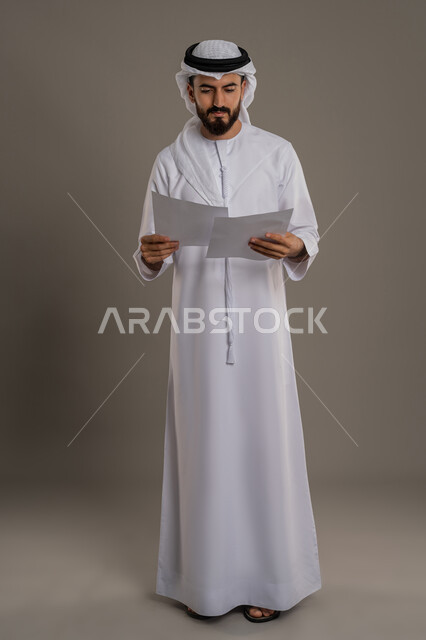 اعداد التقارير الخاصة بالعمل، التدقيق في قراءة الملاحظات، بورتريه لرجل عربي خليجي اماراتي يرتدي الكندورة والغترة ينظر إلى أوراق بيضاء، مراجعة ومتابعة الخطط والمشاريع بإيماءات التركيز والاندماج، الجسم بالطول الكامل، خلفية رمادية