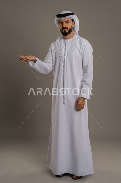 إمساك وإحتواء عنصر هولوجرام، بورتريه لرجل عربي اماراتي خليجي يرتدي الكندورة والغترة يرفع يده لعرض عنصر هولوجرامي ما والنظر إلى الكاميرا بتعابير السرور, ايماءات تدل على السعادة,  الجسم بالطول الكامل، خلفية رمادية