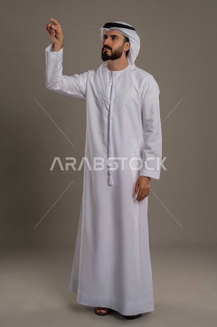 تعابير تدل على التركيز بشيء ما, ايماءات الضغط على عنصر هولوجرام، بورتريه لرجل عربي اماراتي خليجي يرتدي الكندورة والغترة يرفع اصبع السبابة بحركات التفاعل مع عناصر هولوجرامية، الجسم بالطول الكامل، خلفية رمادية