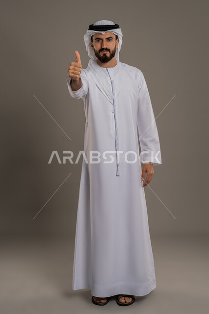 الاهتمام بالأناقة والمظهر الخارجي، مفهوم الوسامة والرجولة والقوة، النظر الى الكاميرا بإيماءات السرور والثقة في النفس، بورتريه لرجل عربي خليجي اماراتي يرتدي الكندورة والغترة  يرفع اصبع الابهام بتعابير الاعجاب والقبول، الجسم الكامل بالطول، خلفية رمادية