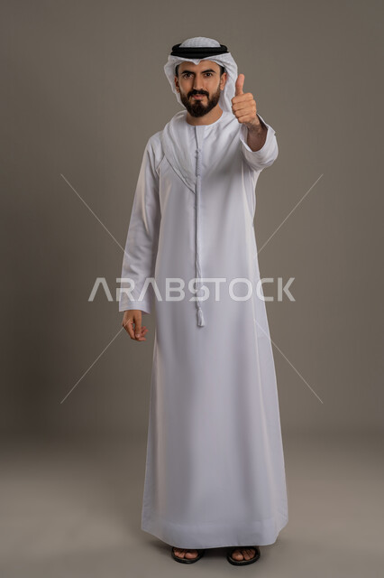 مفهوم الوسامة والرجولة والقوة، النظر الى الكاميرا بإيماءات السرور والثقة في النفس، الاهتمام بالأناقة والمظهر الخارجي، بورتريه  لرجل عربي خليجي اماراتي يرتدي الكندورة والغترة  يرفع اصبع الابهام بتعابير الاعجاب والقبول، الجسم الكامل بالطول، خلفية رمادية