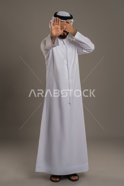 مفهوم الرفض والامتناع والانزعاج، رفع كف اليد بتعابير التوقف وعدم الاقتراب، بورتريه لرجل عربي خليجي اماراتي يرتدي الكندورة والغترة البيضاء يضع يده على عينيه بايماءات الخجل، النهي عن فعل امر ما، الجسم بالطول الكامل، خلفية رمادية