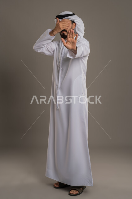 رفع كف اليد بتعابير التوقف وعدم الاقتراب، مفهوم الرفض والامتناع والانزعاج، بورتريه لرجل عربي خليجي اماراتي يرتدي الكندورة والغترة البيضاء يضع يده على عينيه بايماءات الخجل، النهي عن فعل امر ما، الجسم بالطول الكامل، خلفية رمادية