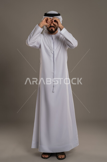 محاولة تقريب الأشياء البعيدة، التخطيط للأهداف والطموحات المستقبلية، حب الاكتشاف والمعرفة والمغامرة، التركيز على شيء ما، بورتريه لرجل عربي خليجي اماراتي يرتدي الكندورة والغترة يضع يديه على عيونه بشكل منظار بإيماءات البحث، صورة الجسم بالطول الكامل، خلفية رمادية