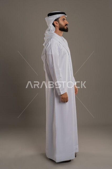 النظر الى مكان ما بإيماءات السرور والثقة في النفس، مفهوم الوسامة والرجولة والقوة، الاهتمام بالأناقة والمظهر الخارجي، بورتريه من الجانب لرجل عربي خليجي اماراتي يرتدي الكندورة والغترة  يقف باستقامة، الجسم الكامل بالطول، خلفية رمادية