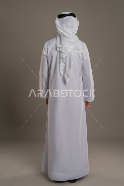 مفهوم الوسامة والرجولة والقوة، الاهتمام بالأناقة والمظهر الخارجي، بورتريه من الخلف لرجل عربي خليجي اماراتي يرتدي الكندورة والغترة  يقف باستقامة، النظر الى مكان ما، الجسم الكامل بالطول، خلفية رمادية