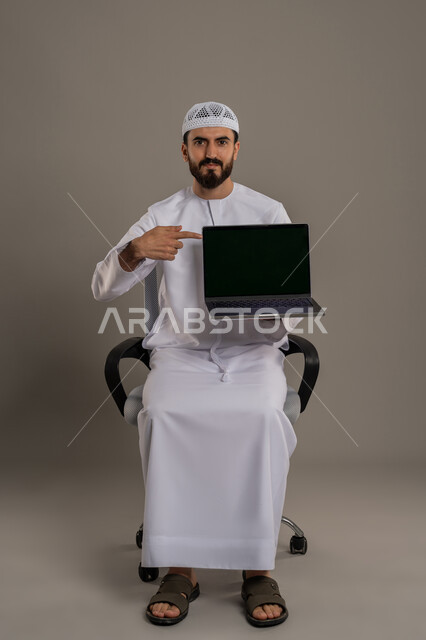 العمل عن بعد، الاشارة باصبع السبابة إلى شاشة سوداء فارغة، تصفح مواقع التواصل الإجتماعي، استخدام جهاز تقني حديث ومتطور لانجاز المشاريع، بورتريه مقرب لرجل عربي خليجي اماراتي يرتدي الكندورة والقبعة البيضاء يجلس على كرسي مريح ويستخدم الحاسوب المحمول، خلفية رمادية