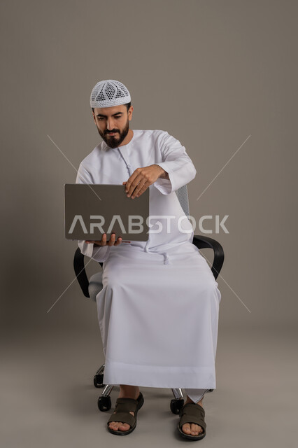 Browsing social media and working remotely, using a modern and advanced technical device to complete projects, close-up portrait of an Arab Gulf Emirati man wearing a kandura and a white hat sitting on a comfortable chair and using a laptop, gray background