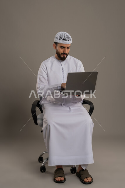 استخدام جهاز تقني حديث ومتطور لانجاز المشاريع، الكتابة على لوحة المفاتيح، تصفح مواقع التواصل الإجتماعي والعمل عن بعد، بورتريه مقرب لرجل عربي خليجي اماراتي يرتدي الكندورة والقبعة البيضاء يجلس على كرسي مريح ويستخدم الحاسوب المحمول، خلفية رمادية