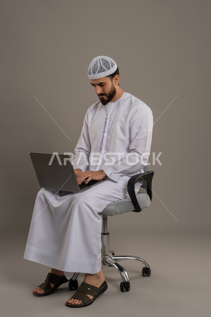 الكتابة على لوحة المفاتيح، استخدام جهاز تقني حديث ومتطور لانجاز المشاريع، تصفح مواقع التواصل الإجتماعي والعمل عن بعد، بورتريه مقرب لرجل عربي خليجي اماراتي يرتدي الكندورة والقبعة البيضاء يجلس على كرسي مريح ويستخدم الحاسوب المحمول، خلفية رمادية