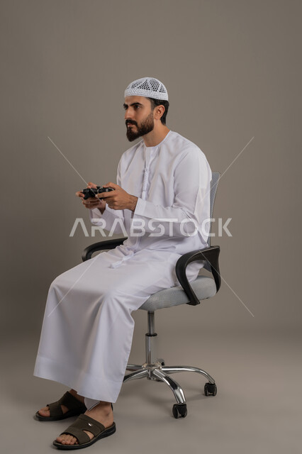 Playing recreational video games, spending free time in a fun activity, using a technological device and modern technology, portrait of an Arab Gulf Emirati man wearing a kandura and a white hat sitting on a comfortable chair holding a joystick in his hands, gray background