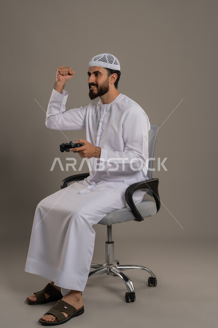 قضاء وقت الفراغ بنشاط ممتع، رفع قبضة اليد بإيماءات الحماس والفوز، استخدام جهاز تكنولوجي وتقنية حديثة، بورتريه لرجل عربي خليجي اماراتي يرتدي الكندورة والقبعة البيضاء يجلس على كرسي مريح يمسك بيديه عصا التحكم، اللعب بألعاب الفيديو الترفيهية, خلفية رمادية