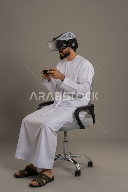جهاز تكنولوجي وتقنية حديثة، بورتريه من الجانب لرجل عربي خليجي إماراتي يرتدي الكندورة والقبعة البيضاء مع نظارة الواقع الإفتراضي يمسك بيده عصا التحكم يجلس على كرسي مريح يقوم باللعب بألعاب الفيديو الترفيهية، دمج العالم الخيالي مع المتعة والتسلية، خلفية رمادية