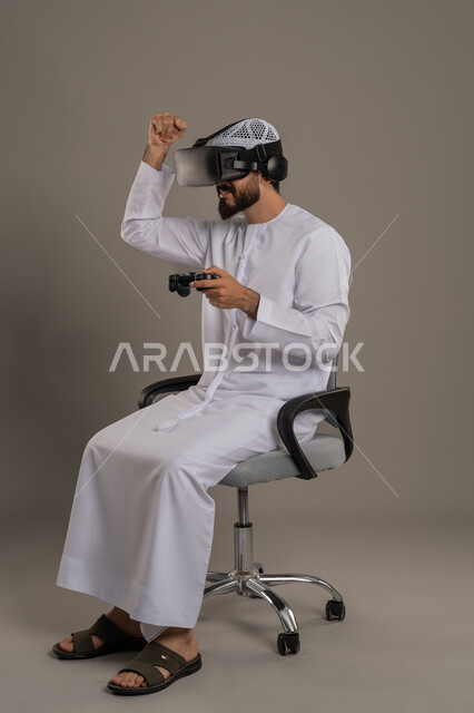Raising fist with enthusiastic gestures, merging fantasy world with fun and entertainment, technological device and modern technology, portrait of an Arab Gulf Emirati man wearing kandora and hat with virtual reality glasses holding joystick in his hand sitting on a comfortable chair playing video games, gray background