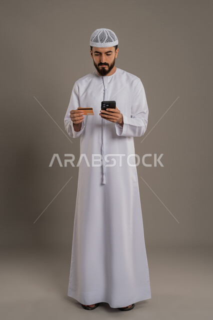 إمكانية الدفع الالكتروني باستخدام البطاقة، النظر إلى الهاتف المحمول بإيماءات التركيز، الشراء والدفع من خلال برامج الجوال، بورتريه لرجل عربي خليجي اماراتي يرتدي الكندورة والقبعة البيضاء يمسك بيده الهاتف المحمول وبطاقة الإئتمان البنكية، خلفية رمادية