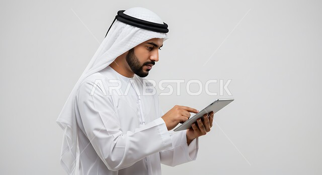 متابعة اخبار وسائل التواصل الاجتماعي من خلال التابلت، الدردشة والتحدث مع الاهل والاصدقاء عبر الانترنت، بورتريه مقرب من الجانب لرجل عربي خليجي اماراتي يرتدي الكندورة والغترة يمسك الجهاز اللوحي، انجاز مهام العمل عن بعد، استخدام جهاز تقني حديث ومتطور، خلفية بيضاء