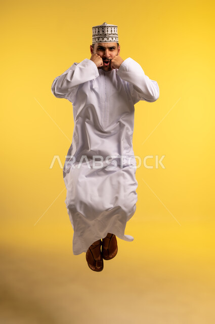 وضع اليدين على الخدين بتعابير الحزن والاستياء، بورتريه لرجل عربي خليجي عماني يرتدي الدشداشة والكمه يقفز عاليا ينظر إلى الكاميرا بإيماءات الغضب، الجسم بالطول الكامل, خلفية صفراء