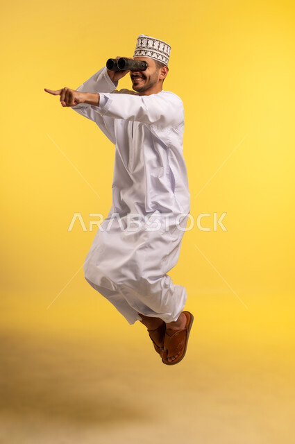 الاشارة باصبع السبابة إلى شيء ما، حب الاستكشاف والمغامرة, رؤية الاشياء البعيدة عبر التلسكوب، التخطيط المستقبلي للأهداف والطموحات، بورتريه لرجل عربي خليجي عماني يرتدي الدشداشة والكمه يقفز عاليا يمسك منظار ثنائي العينين، الجسم بالطول الكامل, خلفية صفراء