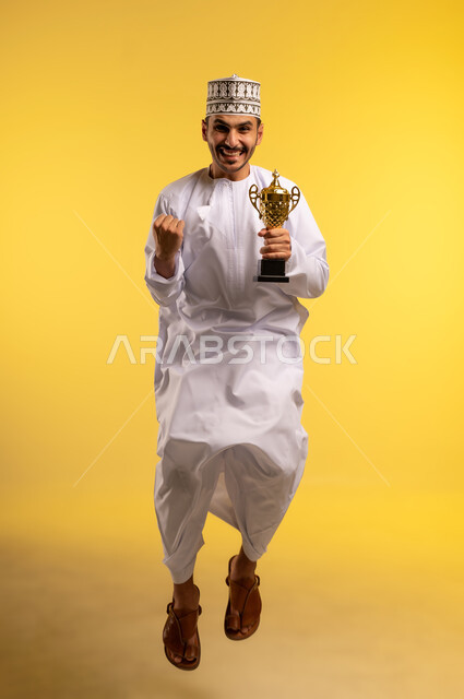 الحصول على المركز الأول في المسابقة، بورتريه لرجل عربي خليجي عماني يرتدي الدشداشه والكمه يقفز عاليا يحمل بيده الكأس الذهبية ينظر إلى الكاميرا بايماءات السرور والحماس، التتويج بالبطولة والفوز، الجسم بالطول الكامل, خلفية صفراء