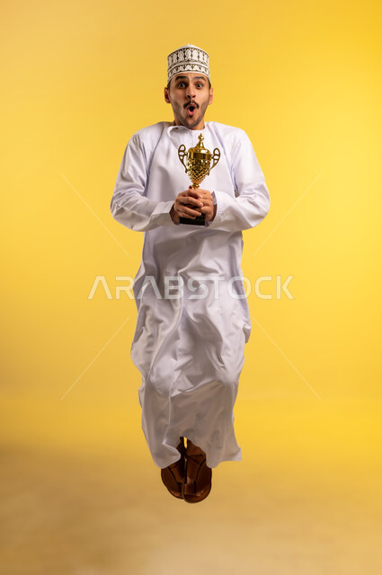 التتويج بالبطولة والفوز، الحصول على المركز الأول في المسابقة، بورتريه لرجل عربي خليجي عماني يرتدي الدشداشه والكمه يقفز عاليا يحمل بيده الكأس الذهبية ينظر إلى الكاميرا بايماءات السرور والحماس، الجسم بالطول الكامل, خلفية صفراء