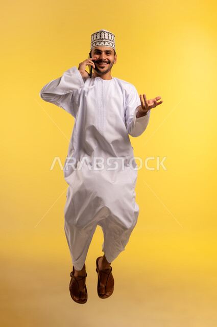 الدردشة والتواصل مع الأهل والاصدقاء عبر الجوال، استخدام جهاز تقني حديث ومتطور، تعابير السرور لسماع اخبار جيدة، بورتريه لرجل عربي خليجي عماني يرتدي الدشداشة والكمة يقفز عاليا يقوم باجراء مكالمة هاتفية عبر الهاتف المحمول، الجسم بالطول الكامل، خلفية صفراء