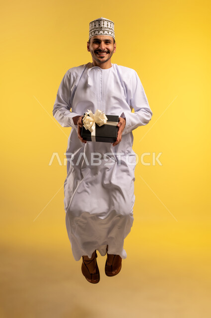 الاحتفال بعيد الميلاد, التعبير عن المحبة والتقدير, بورتريه لشاب عربي خليجي عماني يرتدي الدشداشة والكمة يمسك بيديها علبة هدية يقفز عاليا ينظر إلى الكاميرا بإيماءات السرور والسعادة، تقديم الهدايا في المناسبة السعيدة, الجسم بالطول الكامل، خلفية صفراء