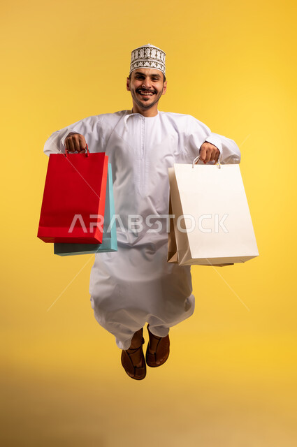 الاستفادة من العروض والتخفيضات الموسميه، الاُثر الايجابي للتسوق، بورتريه لشاب عربي خليجي عماني يرتدي الدشداشة والكمة يحمل بيديه أكياس الشراء يقفز عاليا بإيماءات السرور والسعادة, شراء الهدايا والمستلزمات، الجسم بالطول الكامل، خلفية صفراء