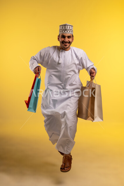 الاستفادة من العروض والتخفيضات الموسميه، الاُثر الايجابي للتسوق، بورتريه لشاب عربي خليجي عماني يرتدي الدشداشة والكمة يحمل بيديه أكياس الشراء يقفز عاليا بإيماءات السرور والسعادة, شراء الهدايا والمستلزمات، الجسم بالطول الكامل، خلفية صفراء