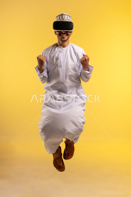 المتعة والتسلية  بالمشاهدة عبر نظارة الواقع الافتراضي, بورتريه لرجل عربي خليجي عماني يرتدي الدشداشة والكمة يقفز عاليا يضع نظارات ثلاثية الأبعاد بتعابير الاندماج والحماس، محاكاة العالم الخيالي، استخدام جهاز تقني حديث ومتطور، الجسم بالطول الكامل، خلفية صفراء