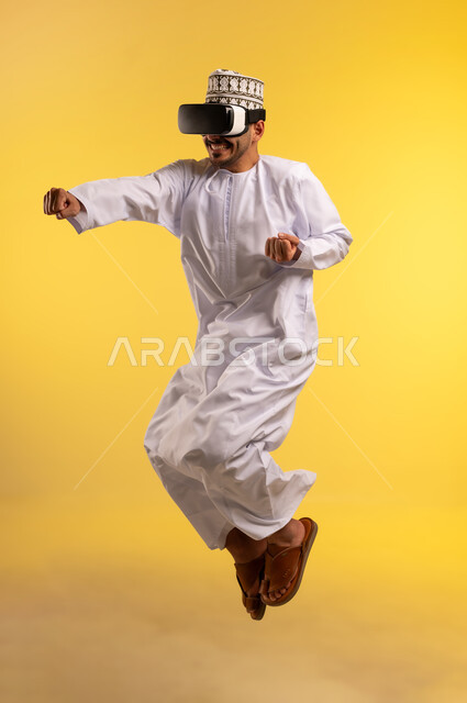 Simulating the imaginary world, fun and entertainment by watching through virtual reality glasses, portrait of an Arab Gulf Omani man wearing a dishdasha and a kummah jumping high wearing 3D glasses with expressions of integration and enthusiasm, using a modern and advanced technical device, full-length body, yellow background