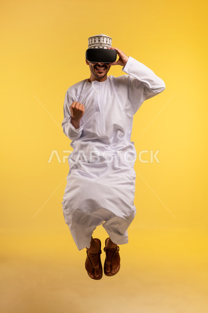 المتعة والتسلية  بالمشاهدة عبر نظارة الواقع الافتراضي, بورتريه لرجل عربي خليجي عماني يرتدي الدشداشة والكمة يقفز عاليا يضع نظارات ثلاثية الأبعاد بتعابير الاندماج والحماس، محاكاة العالم الخيالي، استخدام جهاز تقني حديث ومتطور، الجسم بالطول الكامل، خلفية صفراء