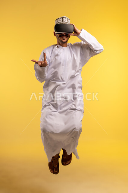 Simulating the imaginary world, fun and entertainment by watching through virtual reality glasses, portrait of an Arab Gulf Omani man wearing a dishdasha and a kummah jumping high wearing 3D glasses with expressions of integration and enthusiasm, using a modern and advanced technical device, full-length body, yellow background