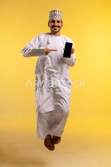 استخدام جهاز تقني حديث ومتطور، النظر إلى الكاميرا بتعابير السرور، عرض تطبيقات وبرامج الجوال، بورتريه لرجل عربي خليجي عماني يرتدي الدشداشة والكمة يقفز عاليا يشير بإصبع السبابة إلى الشاشة السوداء الفارغة عبر الهاتف المحمول، الجسم بالطول الكامل، خلفية صفراء