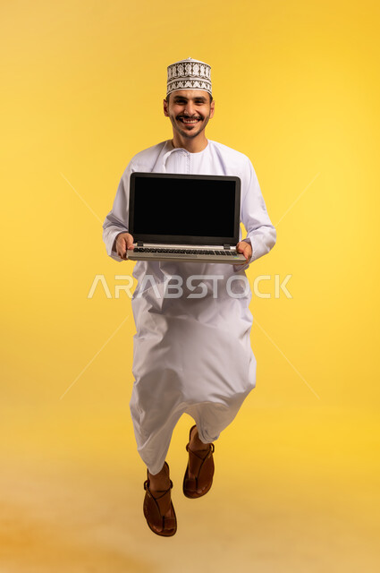 عرض شاشة سوداء فارغة عبر اللابتوب، مفهوم ادارة الأعمال عن بعد، استخدام جهاز تقني حديث ومتطور، بورتريه لرجل عربي خليجي عماني يرتدي الدشداشة والكمة يقفز عاليا يمسك الحاسوب المحمول، دمج التكنولوجيا بالعمل، إنجاز المهام المطلوبة، الجسم بالطول الكامل، خلفية صفراء