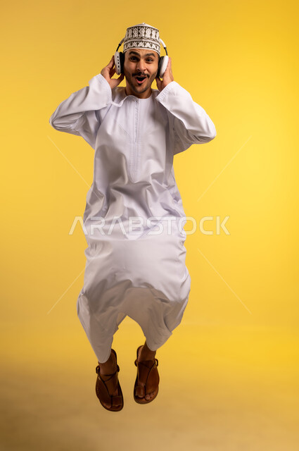مواهب غنائية فنية، الاندماج والتفاعل مع الأنغام الموسيقية، الانصات والاستماع للأغاني باستخدام الهيدفون، بورتريه لرجل عربي خليجي عماني يرتدي الدشداشة والكمة يقفز عاليا يستمع إلى الموسيقى من خلال سماعة الرأس، الجسم بالطول الكامل، خلفية صفراء