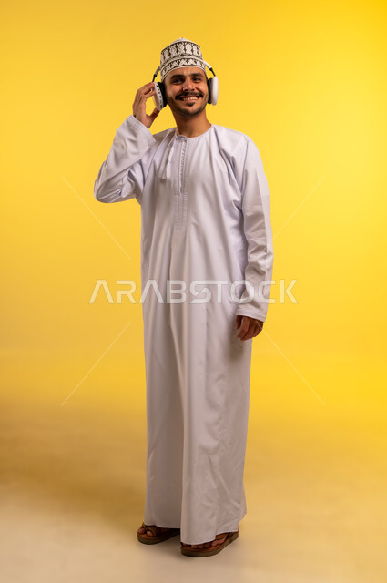 الاندماج والتفاعل مع الأنغام الموسيقية، الانصات والاستماع للأغاني باستخدام الهيدفون، بورتريه لرجل عربي خليجي عماني يرتدي الدشداشة والكمة يستمع إلى الموسيقى من خلال سماعة الرأس، مواهب غنائية فنية، الجسم بالطول الكامل، خلفية صفراء